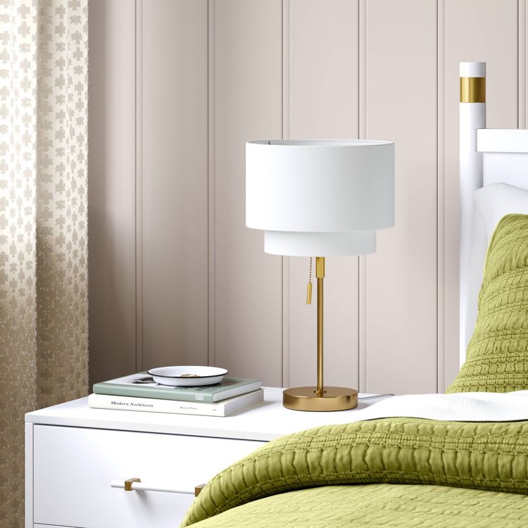 A white colored table lamp with a gold colored base and pull chain sits on a white bedside table next to a green quilted bedspread.