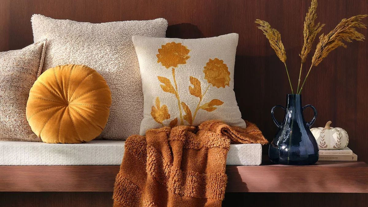 Sunshine floods a room from a nearby window where cream & gold pillows sit on a warm & welcoming padded bench along with a cozy knit blanket. Iconic fall decor including a ceramic white pumpkin & blue vase with greenery sits on the end of the bench.