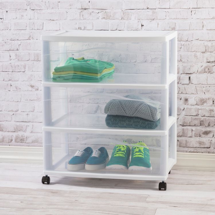 A white utility storage cart with three transparent drawers containing folded clothes and two pairs of shoes, set against a white brick wall.