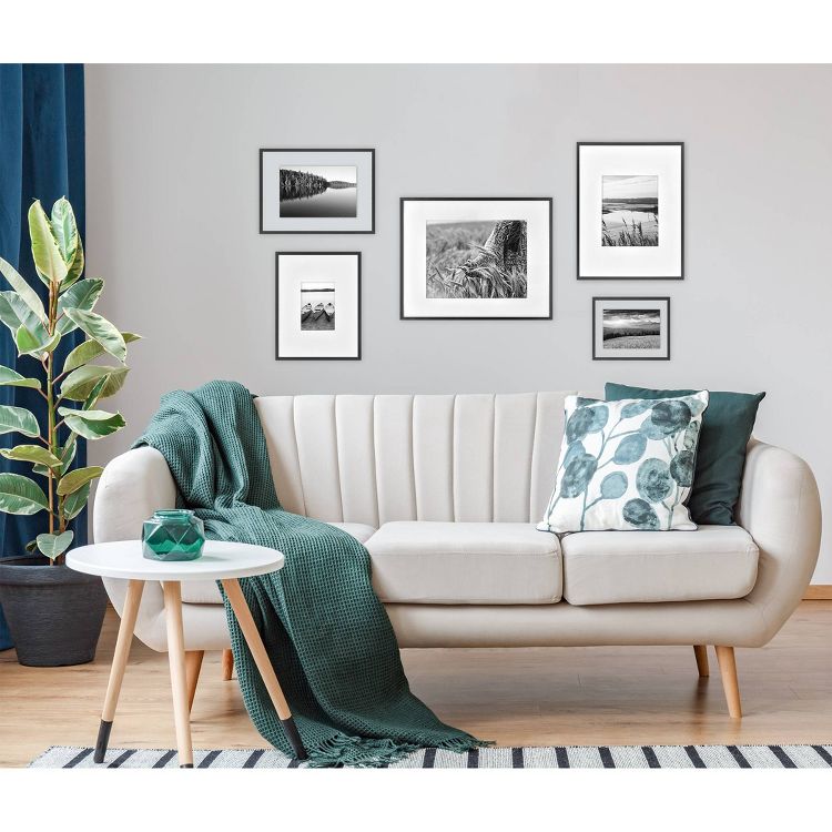 A beige colored sofa with green pillows and a throw is placed in front of a wall with five black and white framed photographs, next to a small round table with a green vase.