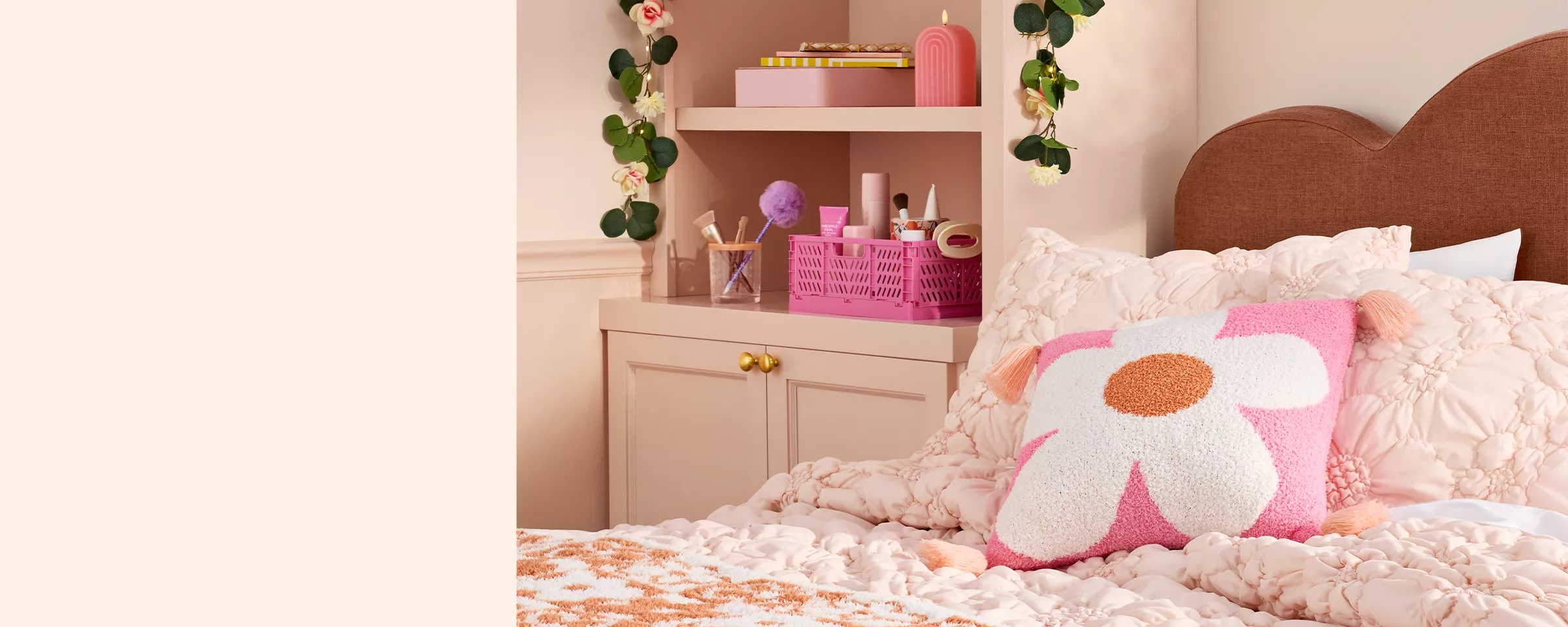 A bright pink, daisy print pillow rests on top a pale pink tufted comforter with floral details. Next to the bed, on a shelf, there’s a bright pink organizer full of beauty items. A floral vine with lights drapes over the shelf creating a soft glow. 