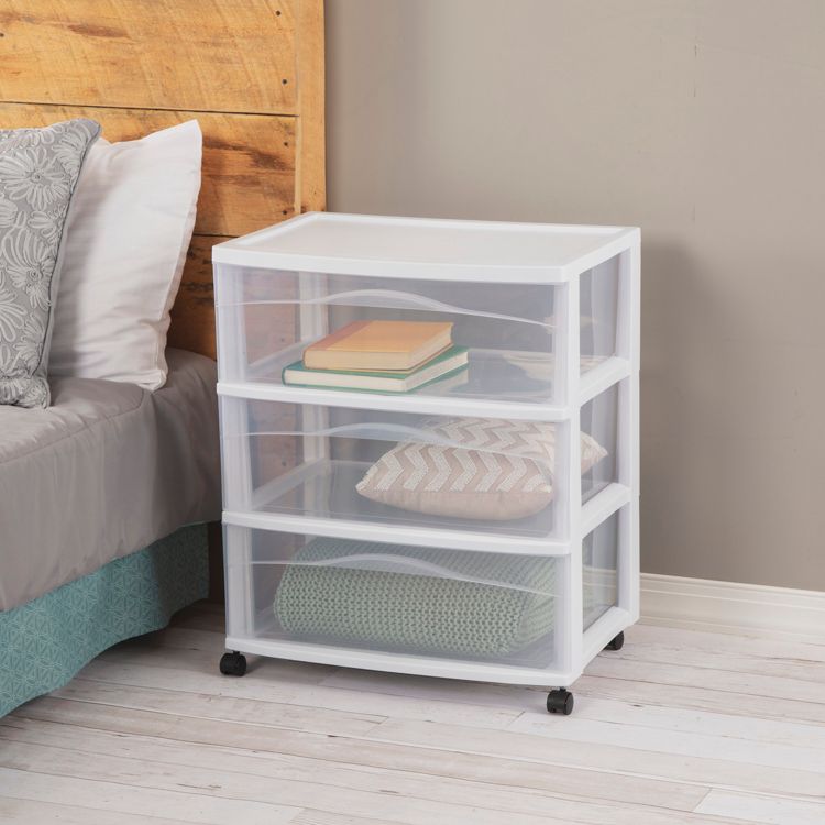 A white colored utility storage cart with three transparent drawers, containing books and folded clothes, placed beside a bed.