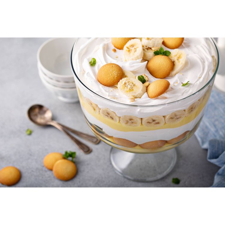 A layered banana pudding dessert in a glass bowl, topped with whipped cream, banana slices, and round cookies, with stacked bowls and spoons in the background.
