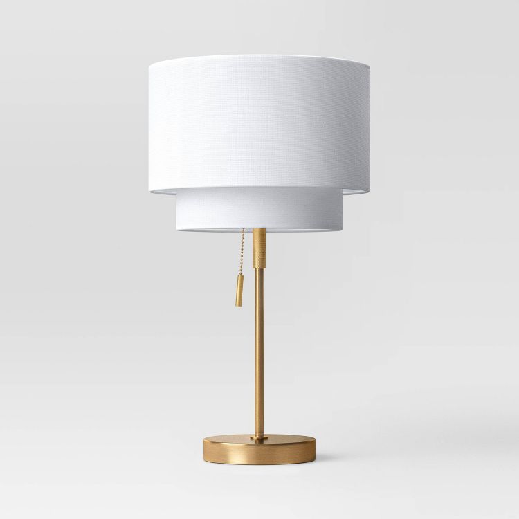 A table lamp with a white colored cylindrical shade and a gold colored base and stand, featuring a pull chain switch.