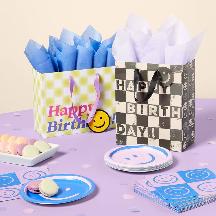 Two gift bags with "Happy Birthday" text, one in a yellow and white checkered pattern with blue tissue paper, and the other in a black and white checkered pattern with white tissue paper, both featuring smiley face tags.