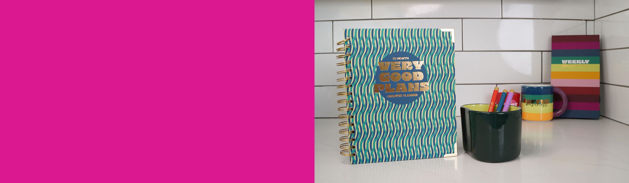 A blue and green planner with a graphic pattern printed with the words "Very good plans" on a counter standing beside mugs, pens, and another striped planner in the background.