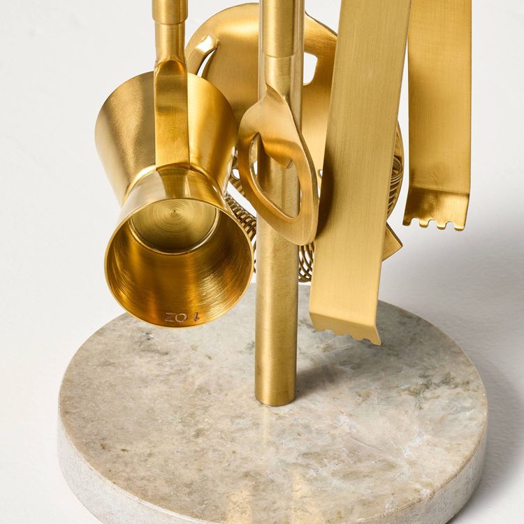 A set of gold colored barware tools, including a jigger marked with "1 OZ," hanging on a stand with a round stone base.