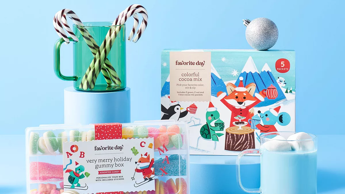 Festive display with holiday treats and drinks on blue stands. A clear box of colorful gummies, a package of colorful cocoa mix with animal illustrations, and a red-striped box of chocolate truffles. Two mugs with creamy drinks and candy canes adorn the scene.