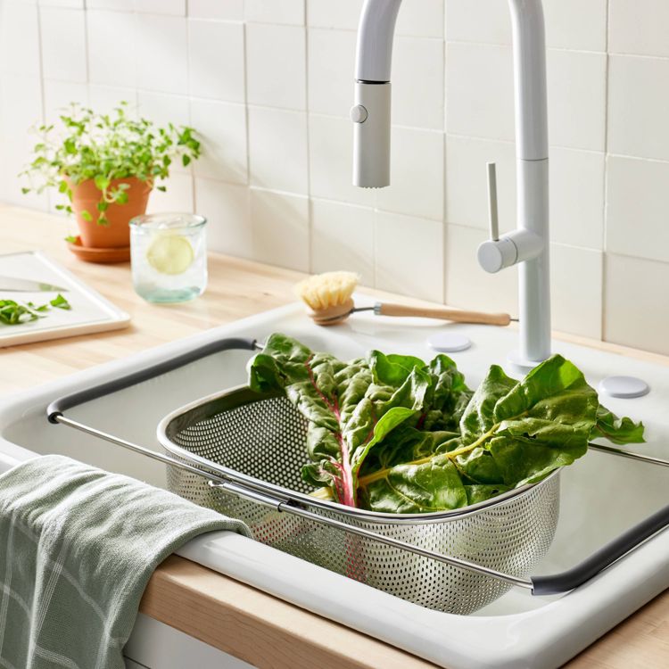 A metal colander with extendable handles is placed over a kitchen sink, holding fresh green leafy vegetables.
