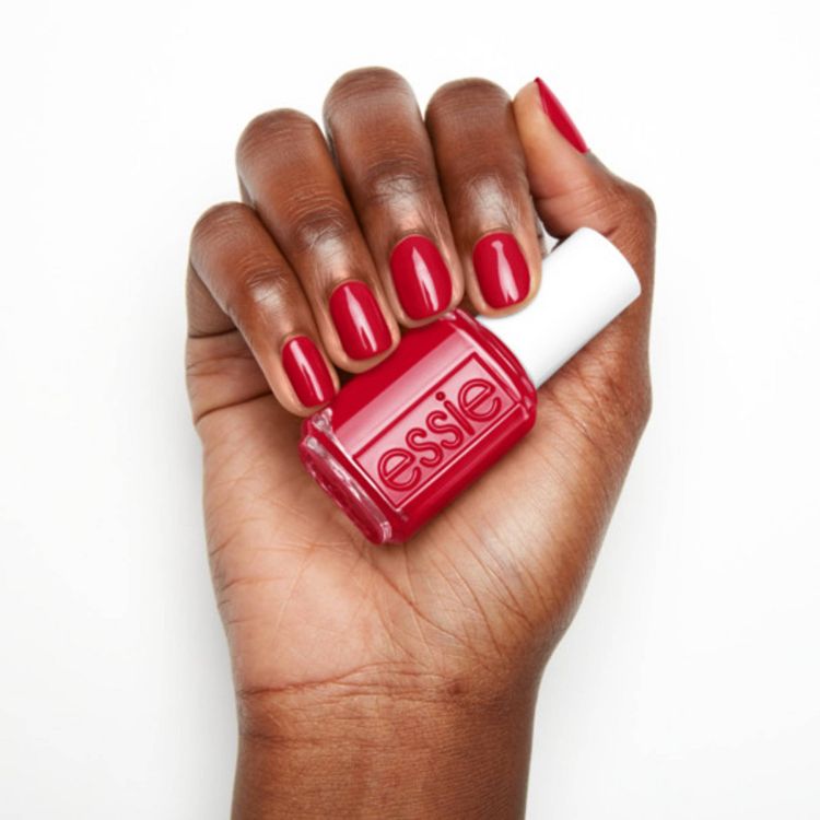 A hand with red colored nails holds a bottle of red colored nail polish with "essie" written on it.