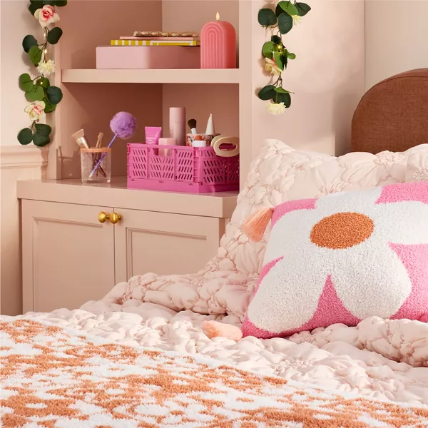 A bright pink, daisy print pillow rests on top a pale pink tufted comforter with floral details. Next to the bed, on a shelf, there’s a bright pink organizer full of beauty items. A floral vine with lights drapes over the shelf creating a soft glow. 