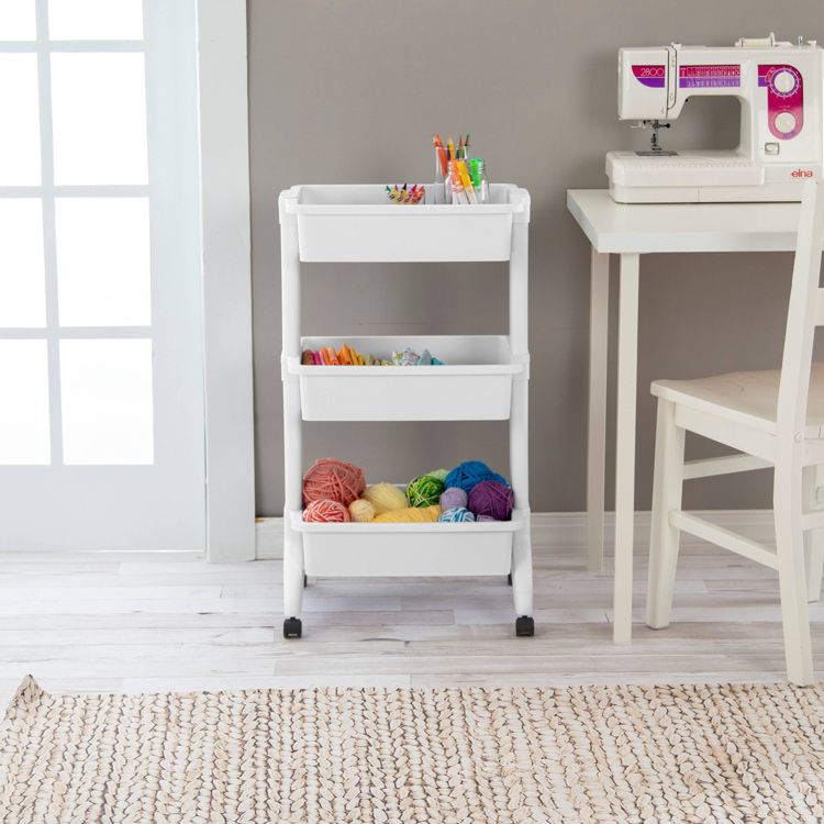 White 3-tier utility cart with wheels, holding colorful yarn balls, pens, and small items, placed on a light wooden floor near a sewing machine and a white chair.