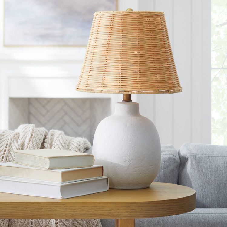 A table lamp with a round white colored base and a woven rattan shade placed on a wooden table.