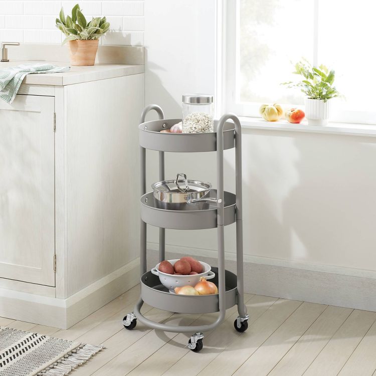 A gray colored three-tier utility storage cart with wheels, holding a jar of grains, a pot, and a bowl of onions, placed in a kitchen setting.