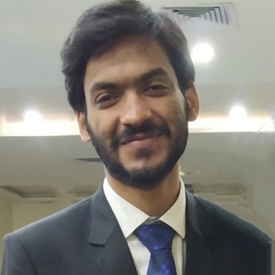 Headshot of Rajshekhar Singhania, a man with short dark hair and a trimmed beard, wearing a black suit, white shirt, and a blue tie. The background is an indoor setting with lights on the ceiling and a reflective surface behind him.
