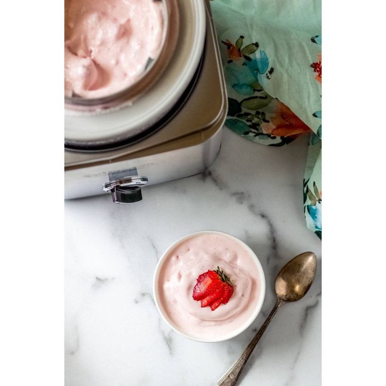 A silver colored ice cream maker with pink ice cream inside, next to a bowl of pink ice cream topped with a strawberry slice and a spoon on a marble surface.