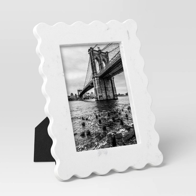 A white colored frame with a wavy edge design holds a black and white photograph of a bridge over water.