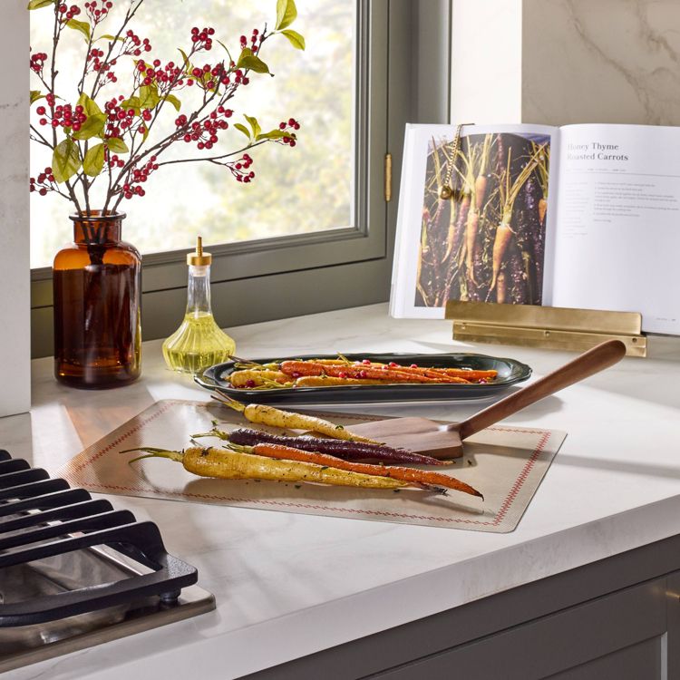 A black colored tray on a kitchen counter holds roasted carrots next to a cookbook and a vase with red berries.
