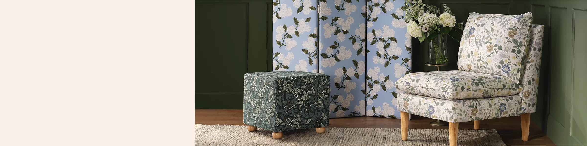 In the corner of a room with deep green walls there’s a white floral accent chair. A light blue room divider with 3 sections & a white hydrangea print stands in front of a wall. There’s a dark green & black ottoman nearby with a leafy design.  