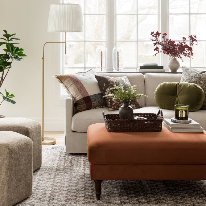 A brightly lit living room with a sofa filled with various pillows, hexagonal ottomans, and an oversized ottoman styled as a coffee table.