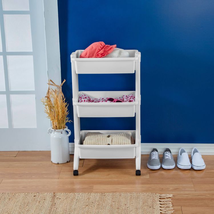 A white three-tiered cart on wheels holding folded clothes, placed against a blue wall.