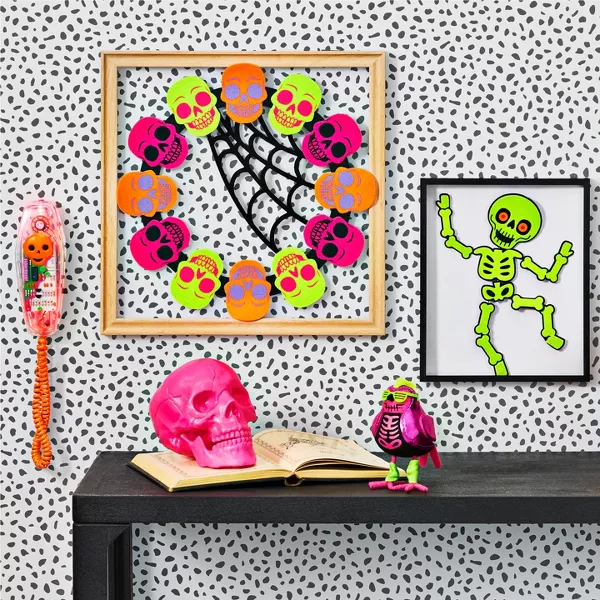 This decor is bright, fun & reflective of the 80s. A pumpkin, pink skull & small purple bird with neon green accessories sit on a side table. A wreath of multi-colored skulls hangs on the wall between a framed skeleton & a Halloween wall phone.