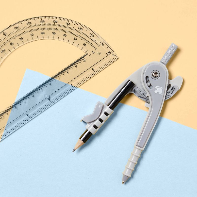 A gray compass and a clear protractor with measurement markings are placed on overlapping sheets of blue and yellow paper.