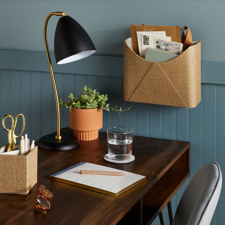 A planner with a beige colored cover and a pen on top is placed on a wooden desk alongside a lamp, a plant, and a glass of water.