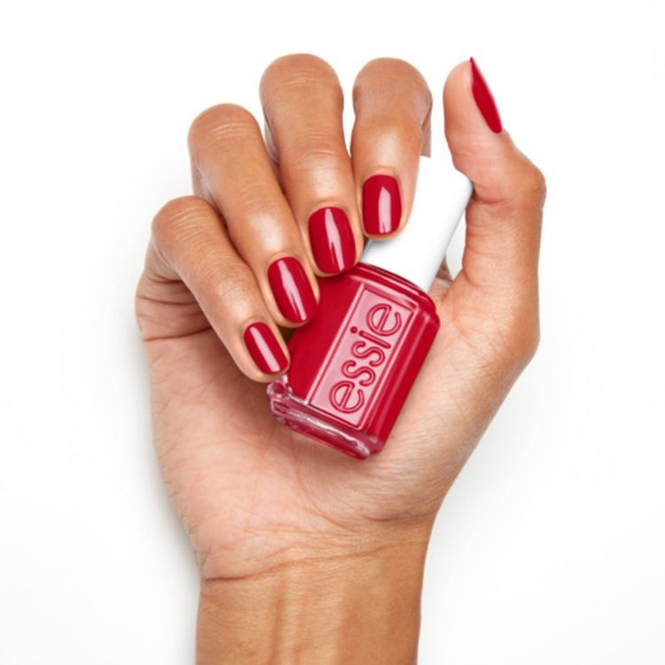 A hand holding a bottle of red colored nail polish with the word "essie" on the bottle, and nails painted in matching red color.