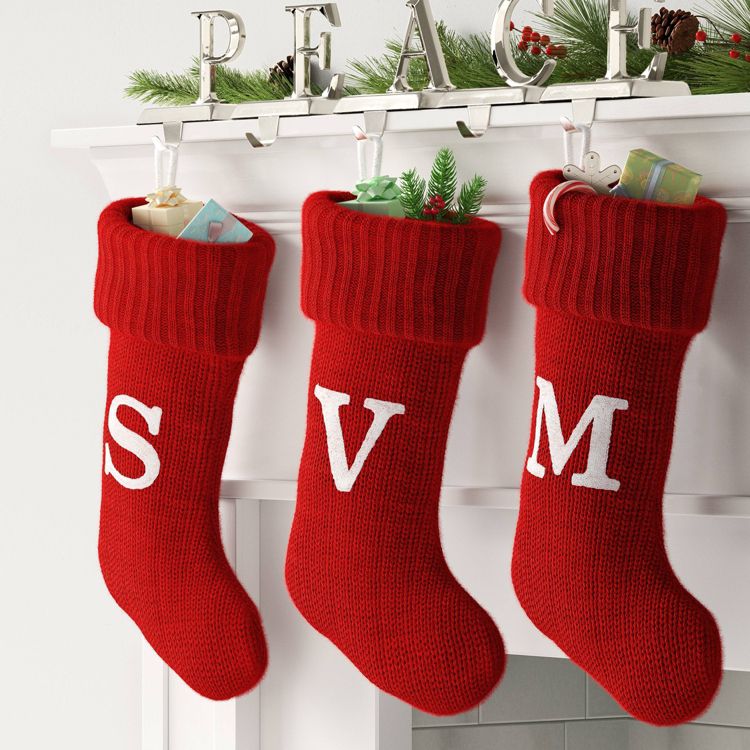 Three red colored stockings with white letters "S," "V," and "M" hang from a mantel, filled with small gifts and greenery.