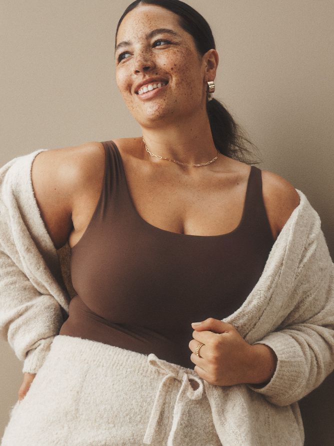 A smiling woman is wearing a casual neutral-beige knit sweater set with a brown tank underneath. The soft cardi sweater is draped over her shoulders. She finishes the look with metallic statement earrings, a delicate necklace and a ring.