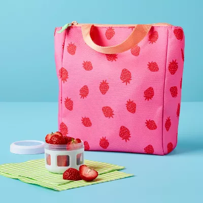 A pink lunch bag decorated with red strawberries on a table next to an open container filled with fresh strawberries, on a green checkered napkin.
