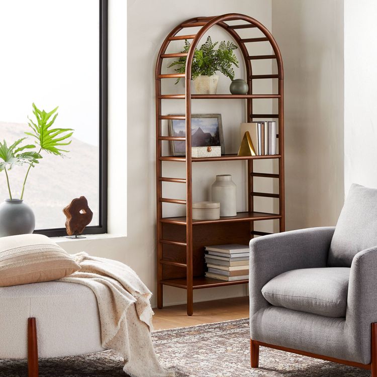 A tall, brown colored arched bookcase with multiple shelves holding books, plants, and decorative items, placed against a wall.