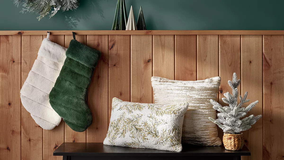 2 white & dark green faux-fur stockings hang on a wooden cabin wall behind a bench. On the bench, 2 white & gold throw pillows rest next to a small, flocked pine tree in a woven basket. This seasonal decor is rustic, cozy & stylish.