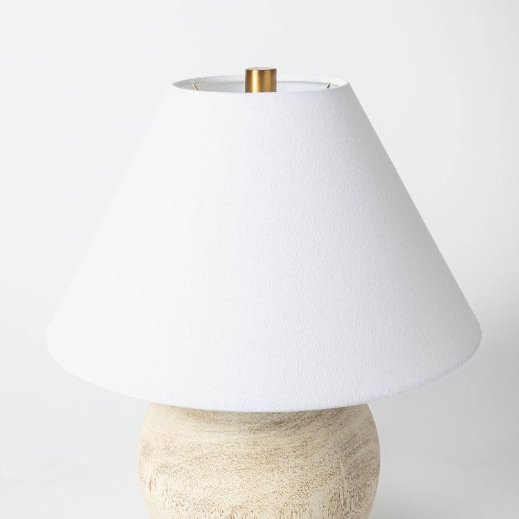 A table lamp with a white colored conical shade and a textured beige colored base.