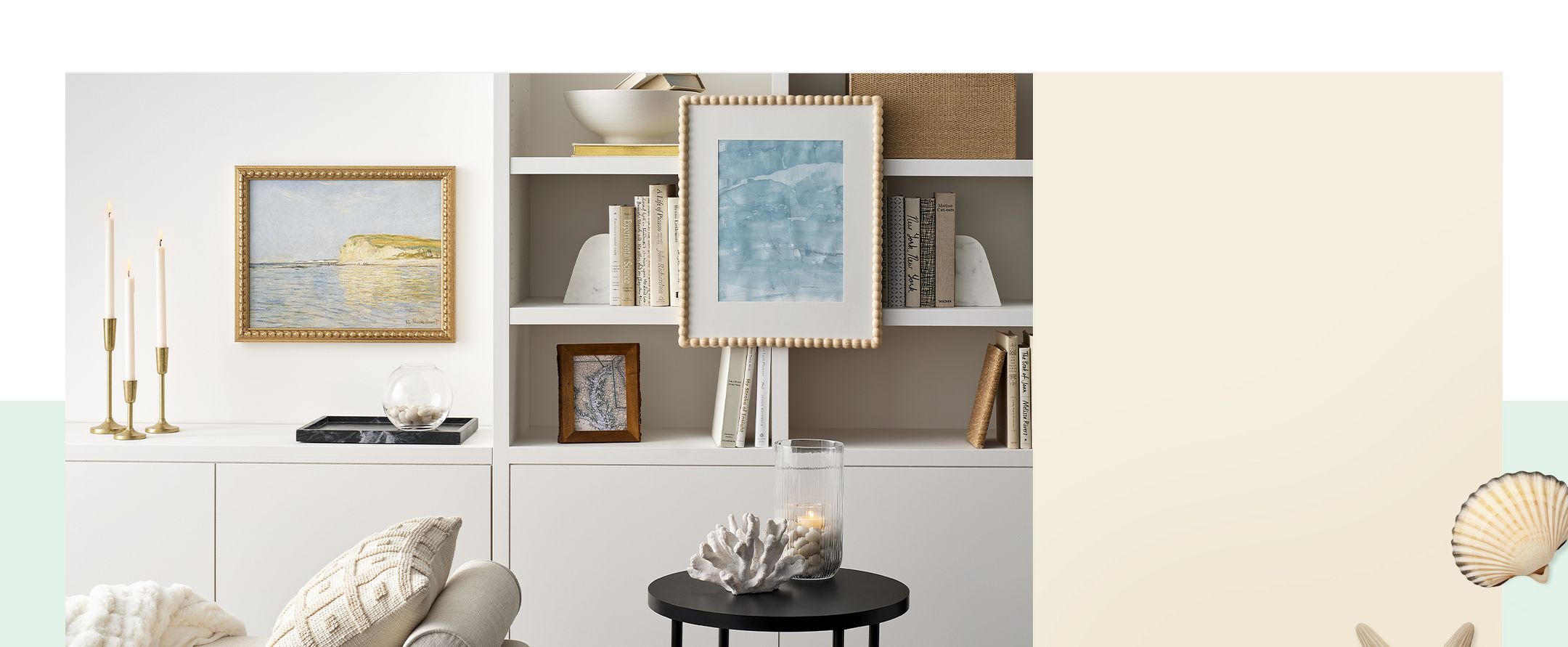 A cozy room with a cushioned bench, pillows, and a soft blanket. Gold-colored candle holders with lit candles sit on a white shelf. Paintings hang above books on a shelf. A small black side table holds a glass candle and coral piece.