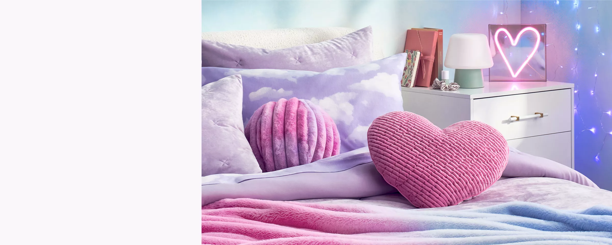 Pink & purple pastel hues bring a peaceful essence to this bedroom. Light purple pillows top the bed with cloud prints, heart shapes & soft velvet textures. Glowing lights hang on the wall & a neon pink heart light rests on the nightstand.   