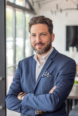 A man with brown hair and beard in a blue suit, white a white collard shirt