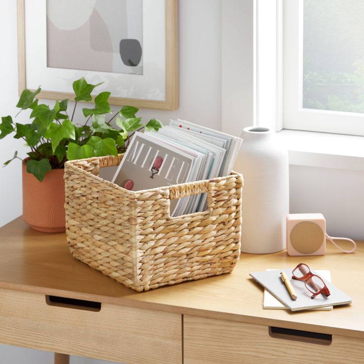 A woven basket holding magazines is placed on a wooden desk next to a potted plant and various desk items.