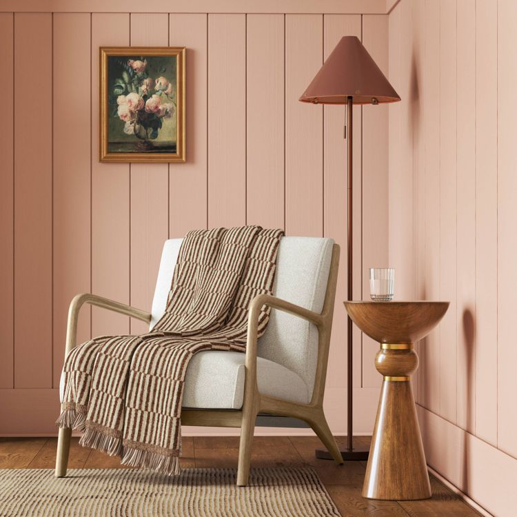 A brown and cream colored striped blanket with fringed edges draped over a light-colored armchair in a room with pink walls.
