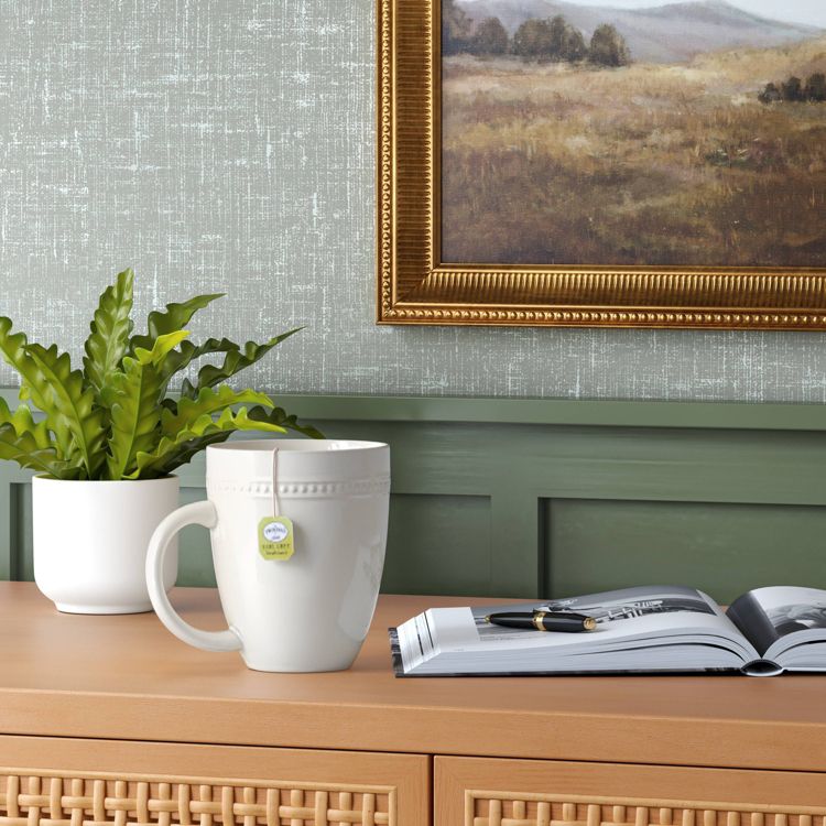 A white colored mug with a tea bag label on the rim is placed on a wooden surface next to a plant and an open book.