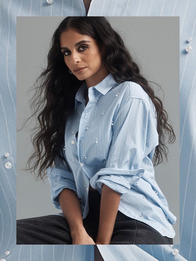 Danie Sierra is wearing a long-sleeve blue button-up collared dress shirt with subtle white stripes. Intricate white beads pop all over the shirt and add a unique three-dimensional modern texture. The sleeves are rolled up and the untucked shirt falls over black dress pants with black sandal heels.