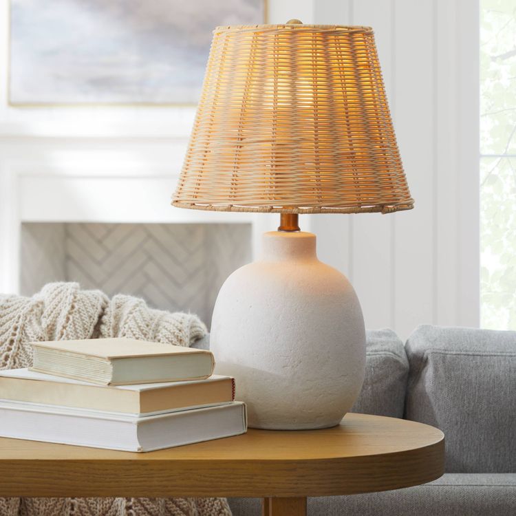 A table lamp with a round white colored base and a woven rattan shade is placed on a wooden table next to a stack of books.