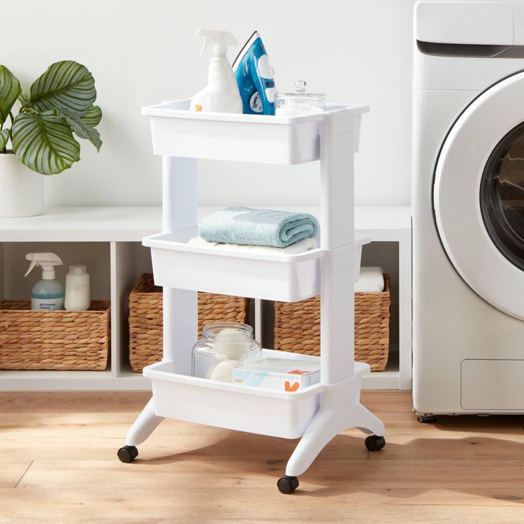 White three-tier utility cart with wheels, holding cleaning supplies, a towel, and other items. It is placed on a wooden floor next to a washing machine, with woven baskets and plants in the background.