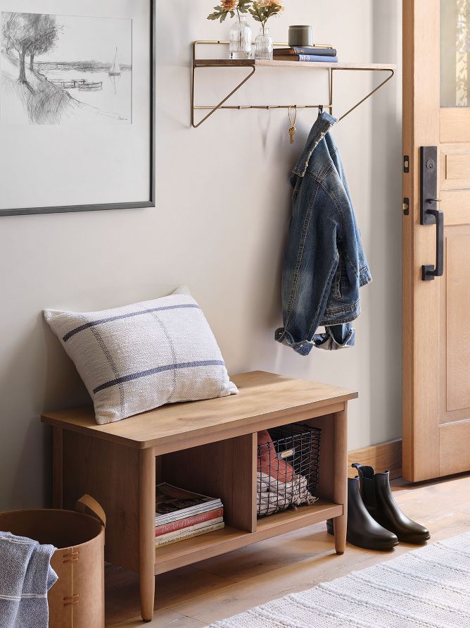 Entry bench with storage sits below a wood and brass wall shelf with hooks holding keys, a jacket and bud vases with faux flowers. 