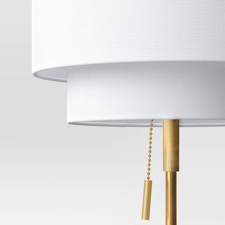 A white colored lampshade with a gold colored pull chain and stand.