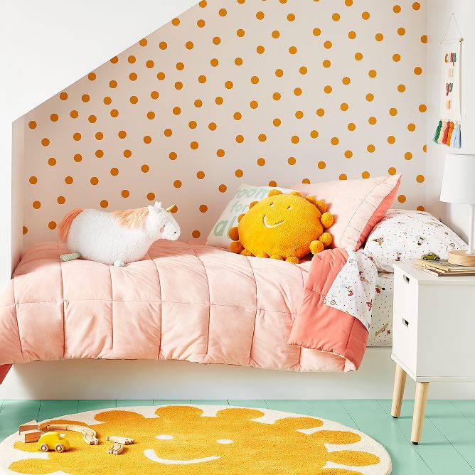 A smiling sun rug sits on a teal floor & warms up the room. A nightstand with a lamp, rests next to a bed nook with a light pink comforter & unicorn plush. Gold dots fill up a wall. A basket of sports balls sits by the bed & an umbrella hangs above. 