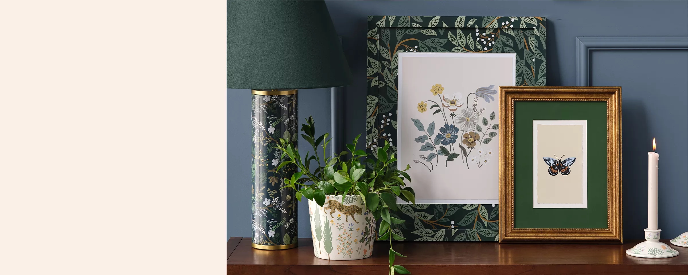 A shelf holds deep blue, black & green décor. There are 2 candlesticks with scalloped holders, a gold framed image of a butterfly, a larger floral framed image, a vine plant growing in a pot with forest imagery & a tall black lamp with floral designs.   