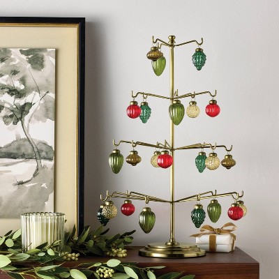 A gold colored ornament stand displaying red, green, and gold colored ornaments, placed on a wooden surface next to a framed painting and greenery.