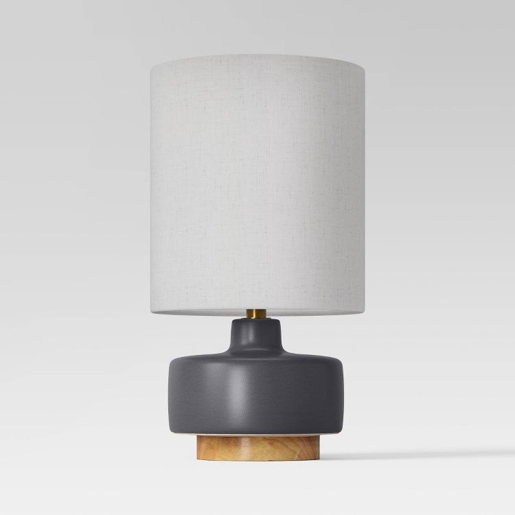 A table lamp with a white colored cylindrical shade, a dark gray colored base, and a wooden platform.